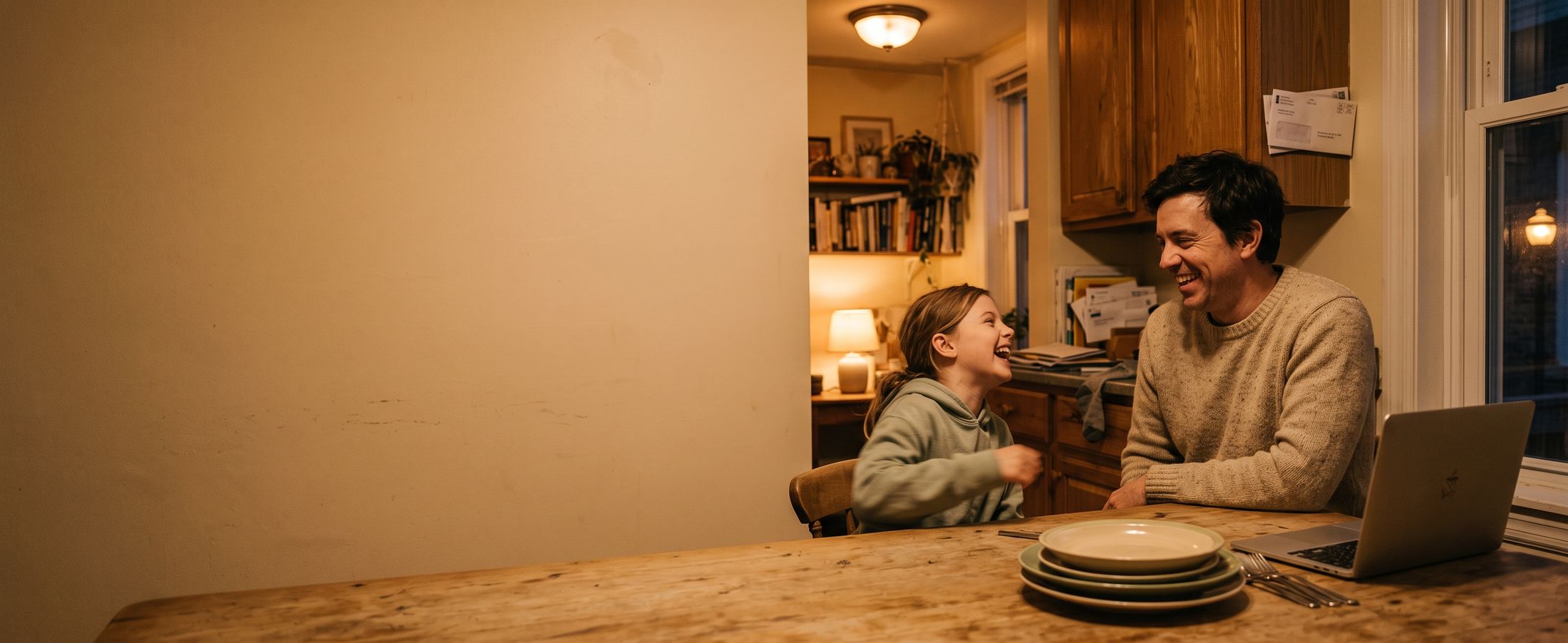 The same kitchen table in the evening. The laptop is closed and pushed aside; a parent and child are laughing together.