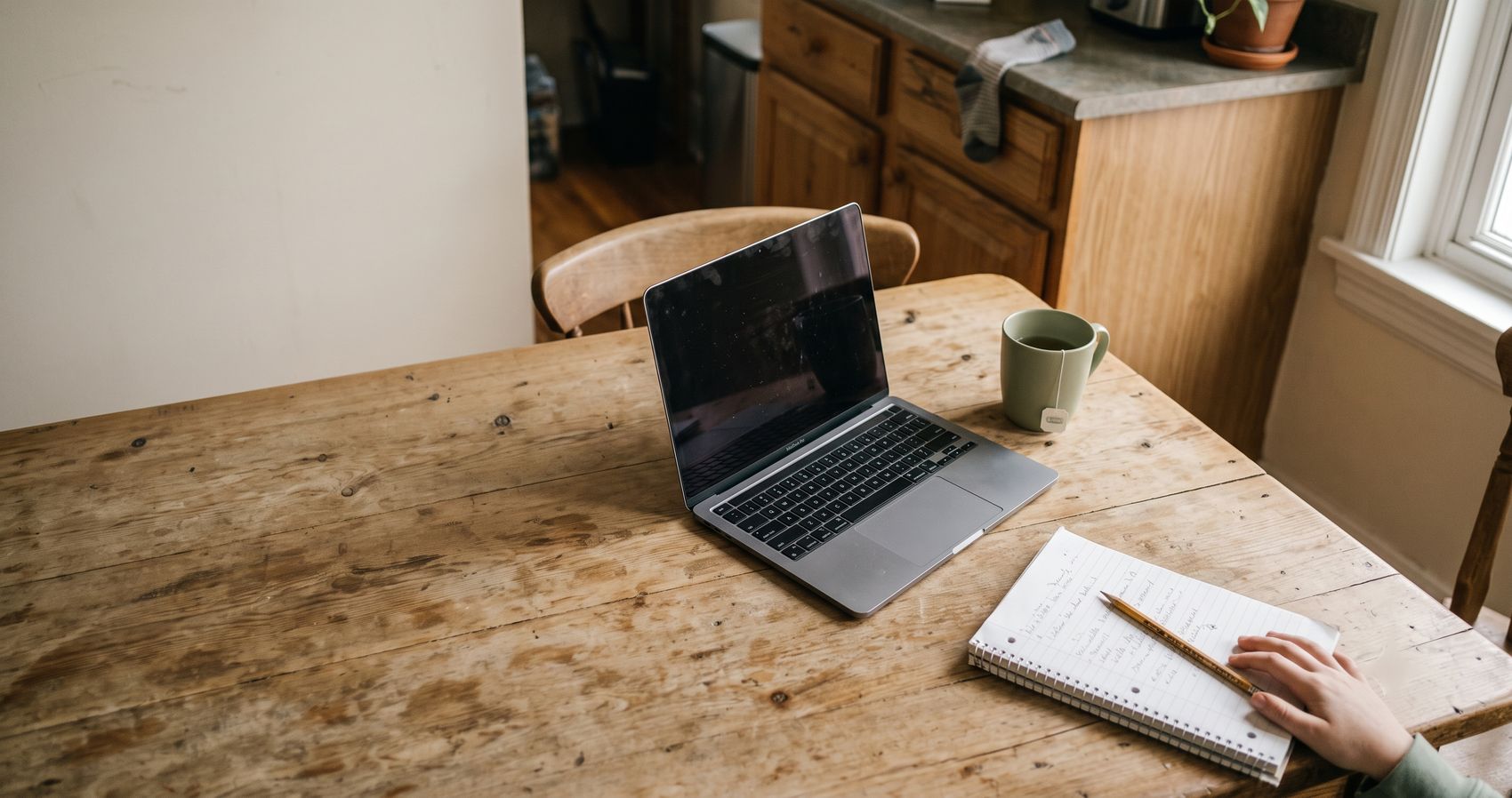 An overhead view of a kitchen table: a closed laptop, a mug, and a spiral notebook with a pencil across it.